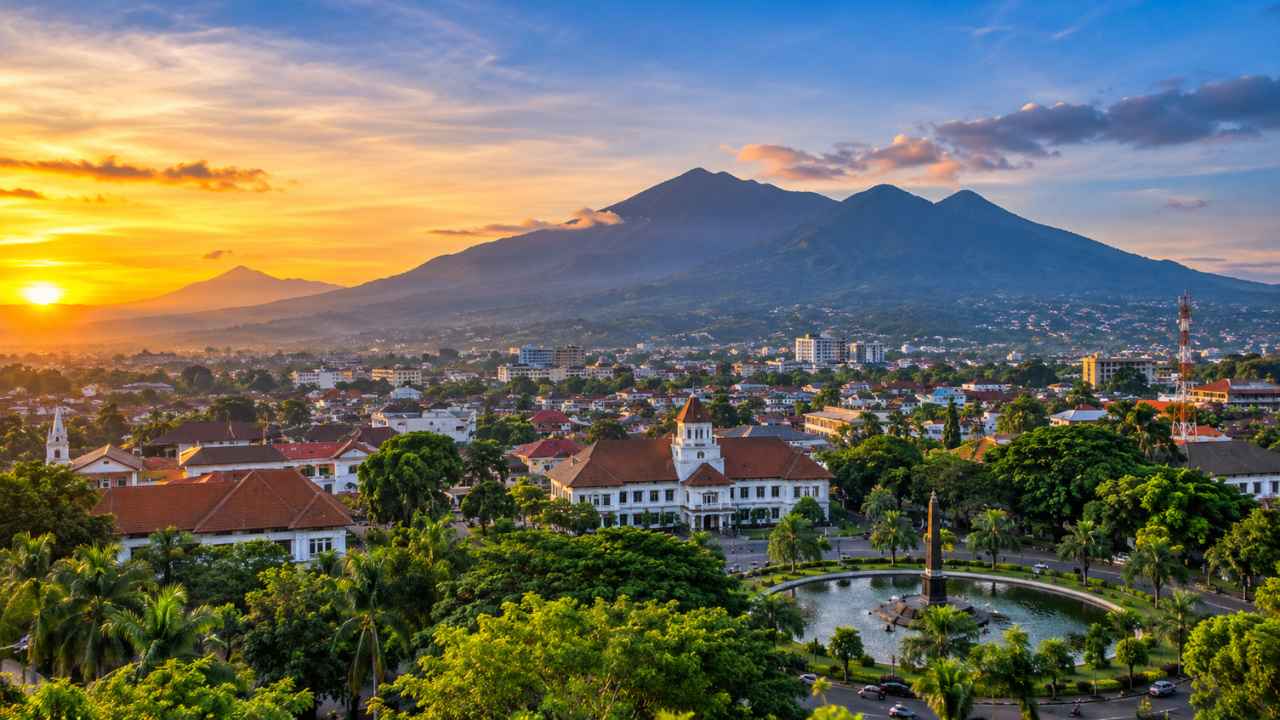 Panorama Kota Malang saat matahari terbenam: gedung Balai Kota beratap merah, bundaran Tugu dengan air mancur, deretan pohon yang rimbun, dan siluet pegunungan di kejauhan