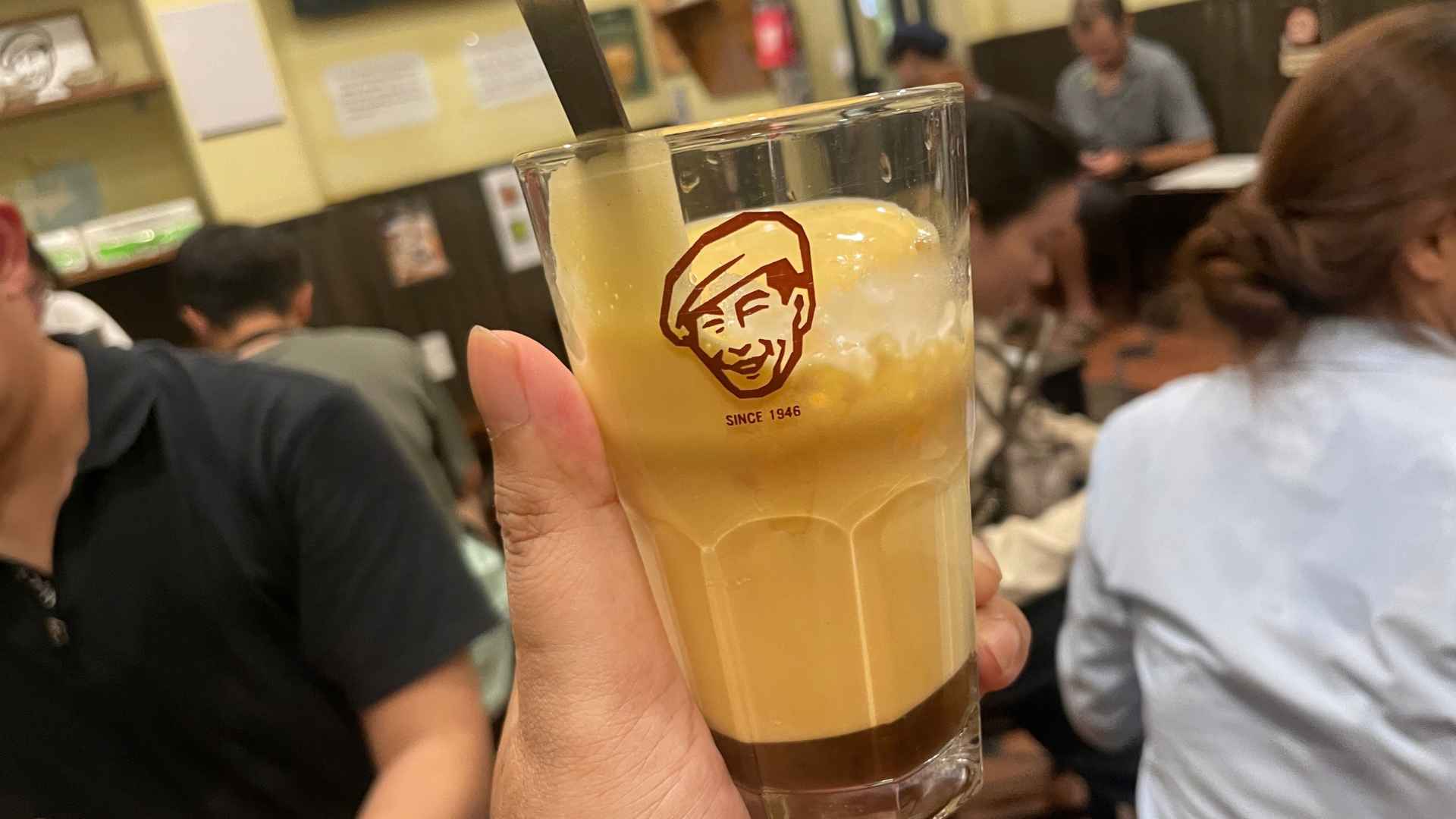 Tangan memegang segelas egg coffee dingin dalam gelas berlogo Café Giảng bertulisan Since 1946 — lapisan kopi hitam pekat di dasar gelas dengan buih krim telur kuning pucat di atasnya