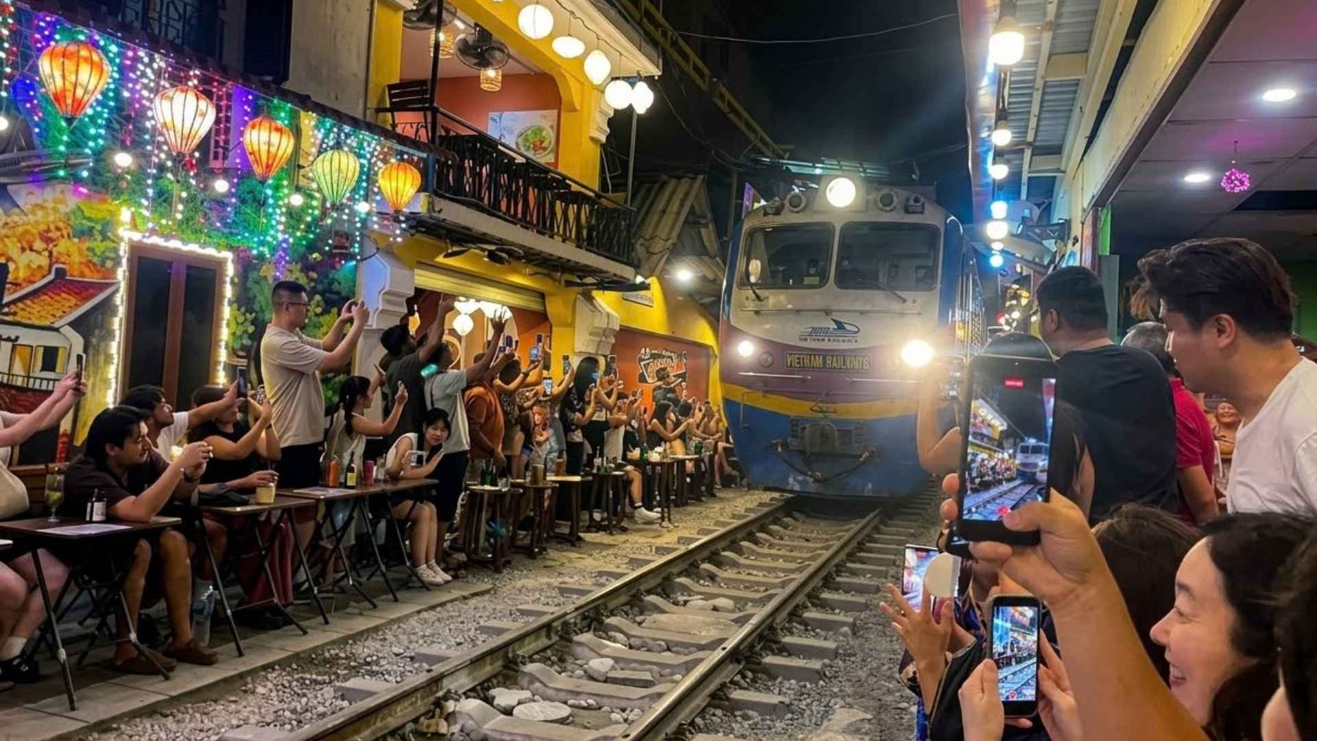 Lokomotif Vietnam Railways dengan lampu menyala melintas pelan di antara kerumunan pengunjung yang duduk di meja kafe tepi rel pada malam hari
