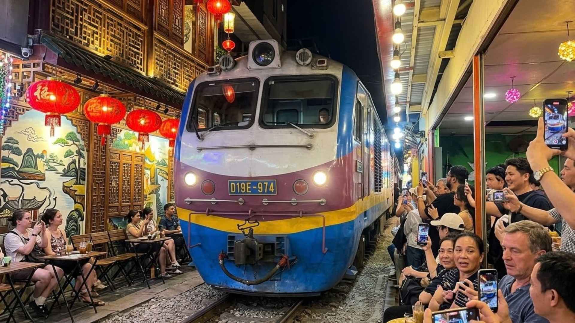 Tampak depan lokomotif Vietnam Railways bertulisan D19E-974 dengan lampu utama menyala, melintas di lorong Train Street yang diapit meja kafe dan lentera merah